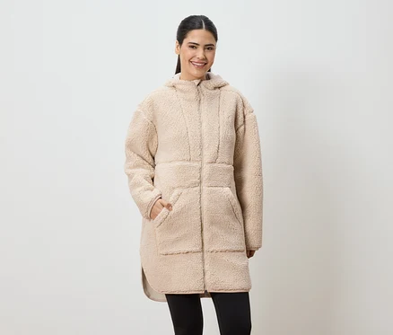 Frau mit Pferdeschwanz trägt einen langen, beigen Teddyfellmantel mit Kapuze und schwarzen Leggings.