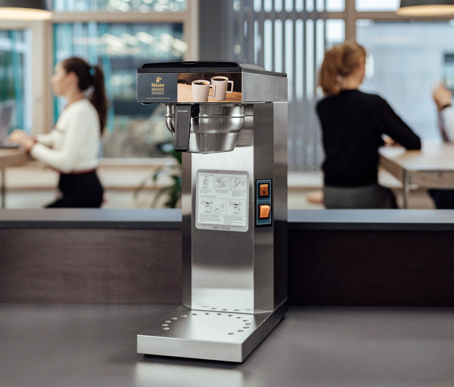 Im Vordergrund befindet sich eine Tchibo Businessline Profi-Filterkaffeemaschine ohne Kanne, im Hintergrund sitzen Leute an Tischen.