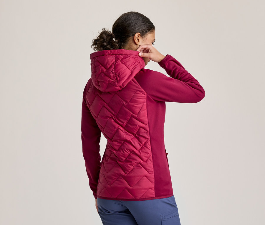 Frau von hinten gesehen, die die Kapuze ihrer dunkelrosa Jacke zurechtrückt. Die Jacke hat einen gesteppten Rücken und eine Kapuze, mit Ärmeln und Seiten aus festem Stoff. Sie trägt blaue Hosen.