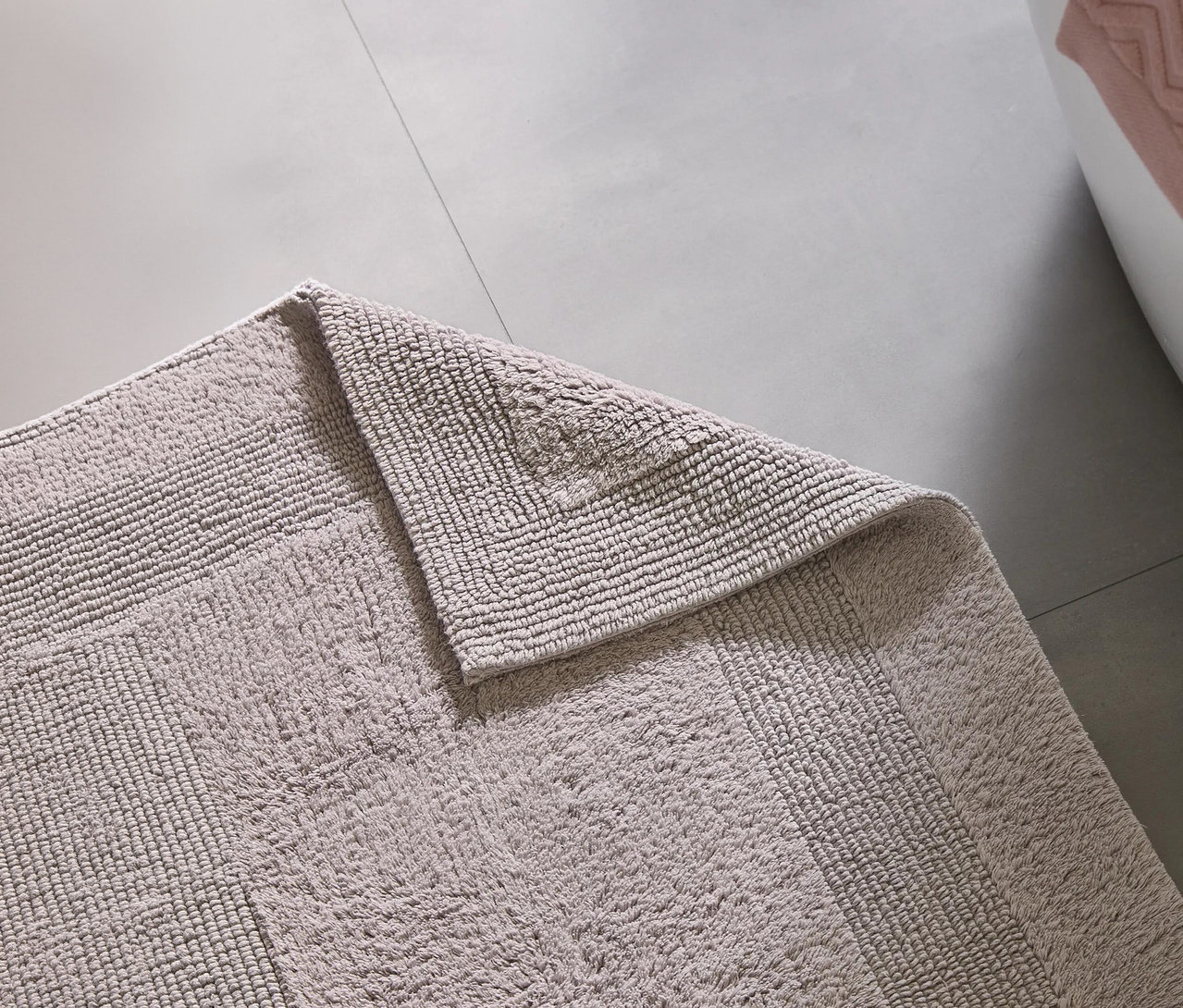 Helle Badematte auf grauem Fliesenboden, eine Ecke ist umgeschlagen. Im Hintergrund sind eine Badewanne und ein rosa Handtuch zu sehen.