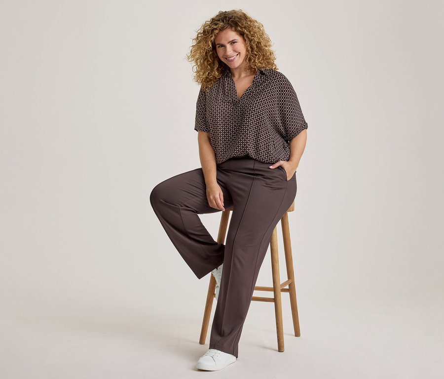 Lächelnde Frau mit lockigem Haar, die ein dunkles gemustertes Kurzarmhemd und eine dunkelbraune Hose trägt, sitzt auf einem Holzhocker, eine Hand in der Tasche und weiße Sneaker an den Füßen.