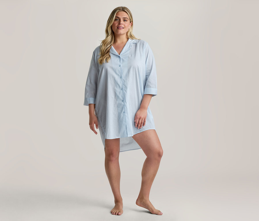 Blonde Frau lächelt, steht barfuß und trägt ein hellblaues Web-Nachthemd mit Kragen, Dreiviertelärmeln und einem High-Low-Saum.