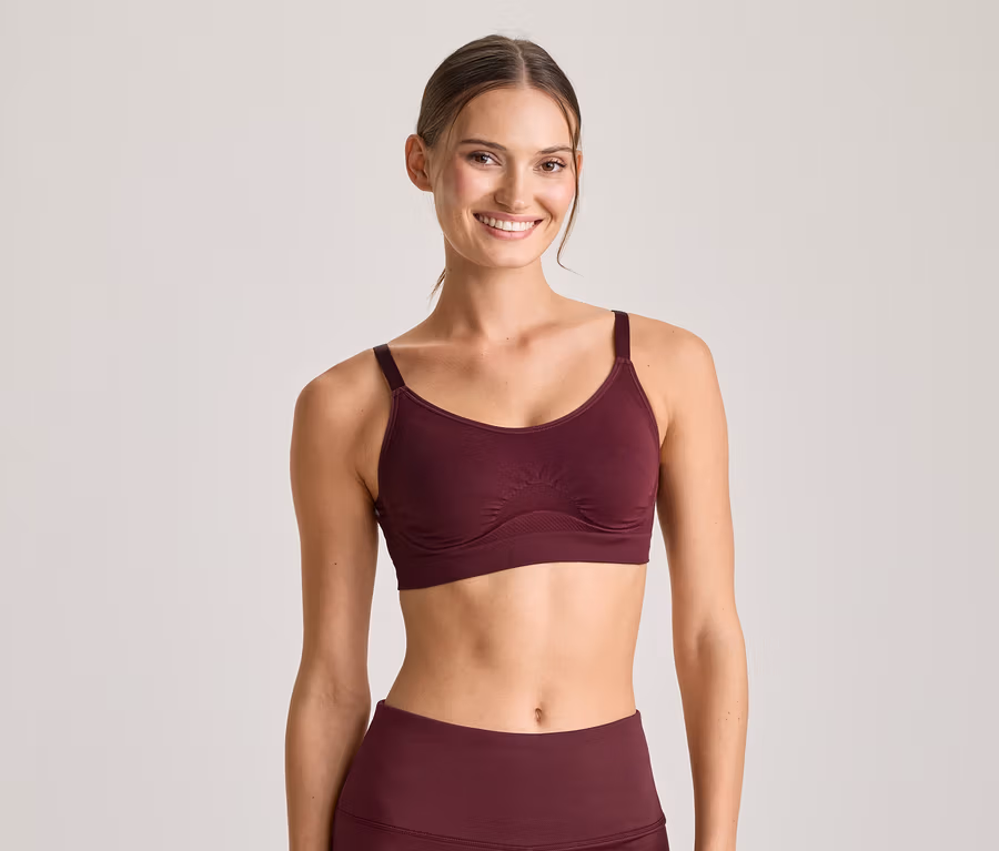 Lächelnde Frau in einem bordeauxroten Sport-BH und passenden Leggings, blickt in die Kamera.