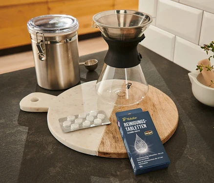 Auf einem Brett liegen durgol® Reinigungstabletten für Kaffeeöle, ein Behälter, ein Kaffeefilter und ein Messlöffel.