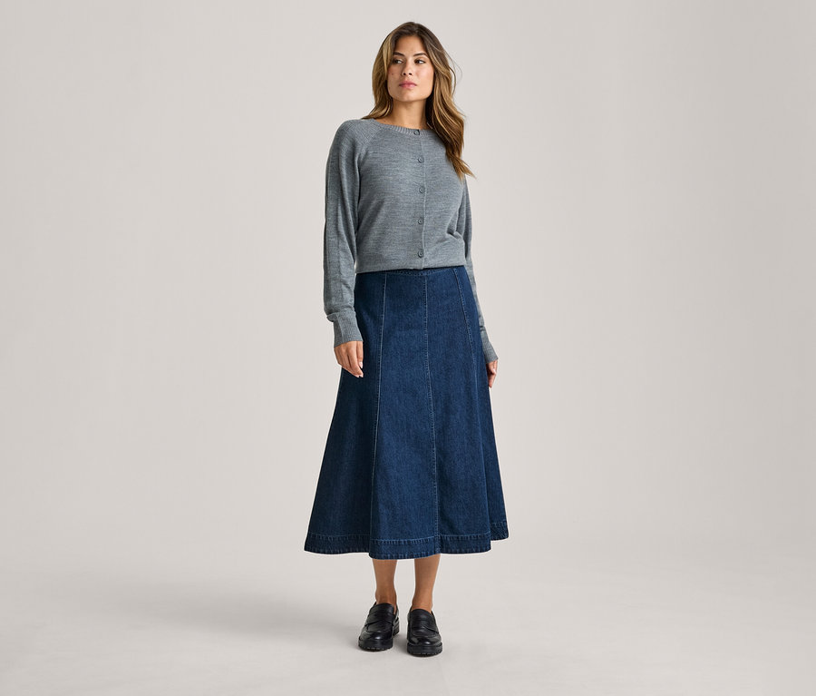 Eine Frau steht da und trägt einen grauen Strickcardigan, einen dunkelblauen Jeans-Midi-Rock in A-Linie und schwarze Loafer.