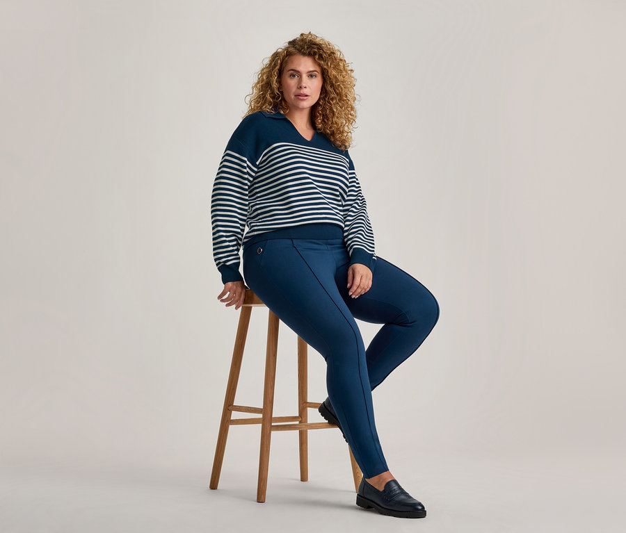 Eine Frau mit lockigem braunem Haar sitzt auf einem Holzschemel und blickt nach vorne. Sie trägt einen marineblau-weiß gestreiften V-Ausschnitt-Pullover, marineblaue schmale Hosen und schwarze Loafer.