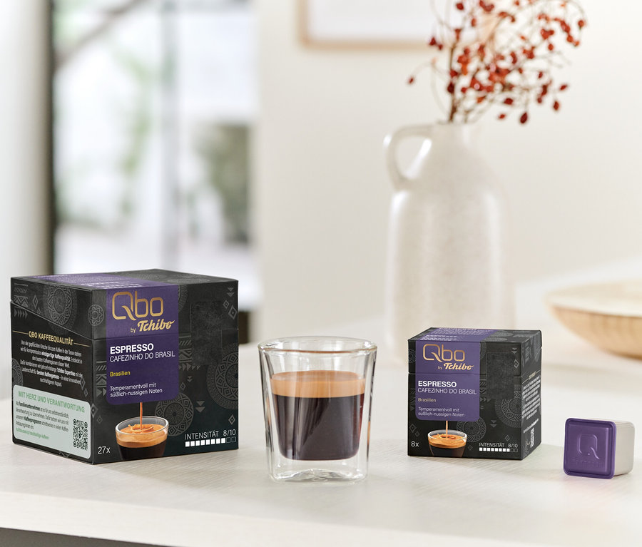 Zwei schwarz-violette Qbo Espresso CAFEZINHO DO BRASIL Kapselpackungen (27 und 8 Stück), ein Glas Espresso und eine einzelne violette Kapsel stehen auf einem weißen Tisch. Im Hintergrund steht eine weiße Vase mit roten Beeren.