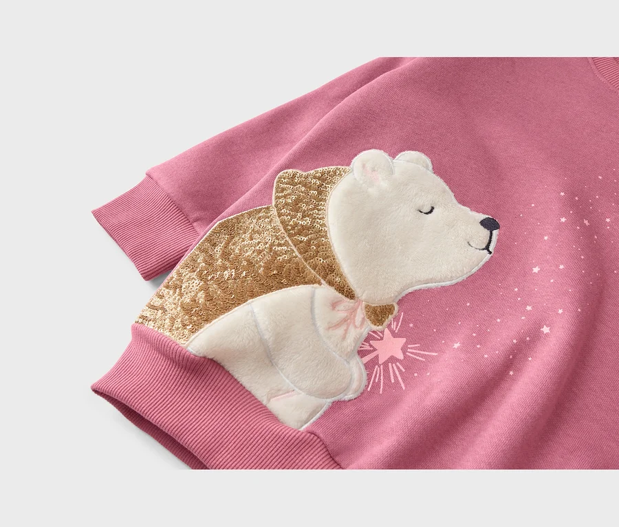 Nahaufnahme eines pinkfarbenen Sweatshirts mit einer Applikation eines weißen Bären. Der Bär trägt einen goldenen Paillettenschal und eine rosa Schleife, umgeben von kleinen weißen Sternen.