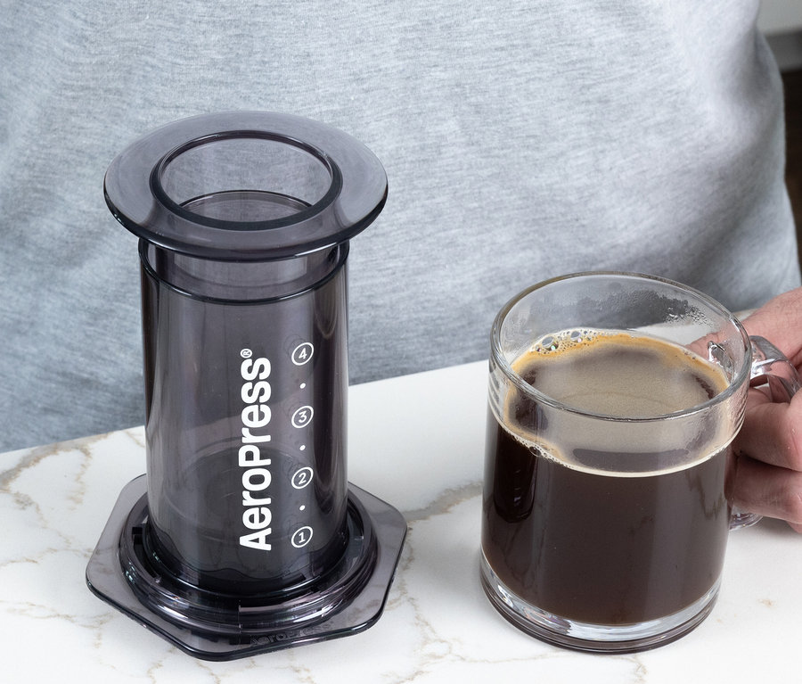 Eine Person hält eine Tasse Kaffee neben einem schwarzen AeroPress Coffee Maker »Clear«.