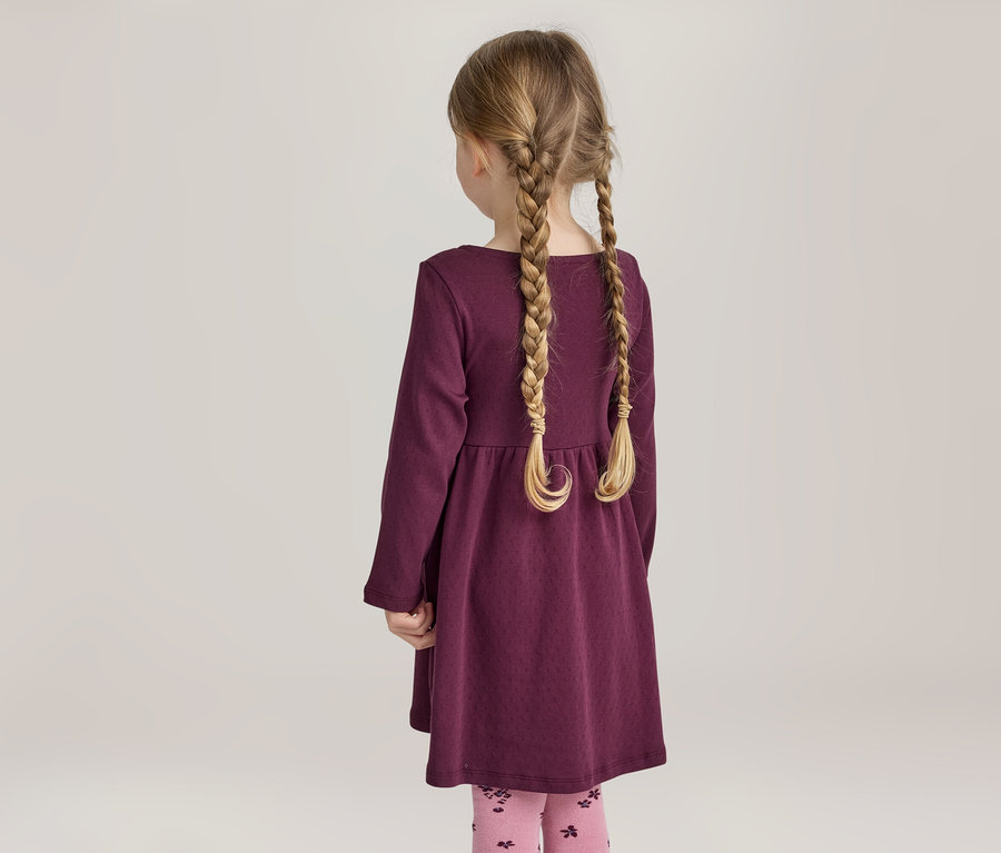 Mädchen von hinten mit blonden Zöpfen, das ein lila Kleid und rosa Leggings mit Muster trägt.