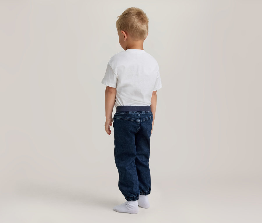 Kleiner Junge mit hellem Haar steht mit dem Rücken zur Kamera, trägt ein weißes T-Shirt, dunkelblaue Jogginghose und weiße Socken.