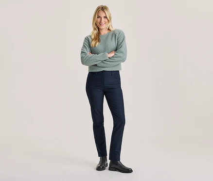 Lächelnde Frau mit blonden Haaren, verschränkte Arme, in hellgrünem flauschigem Pullover, dunkelblauer Hose und schwarzen Stiefeln.