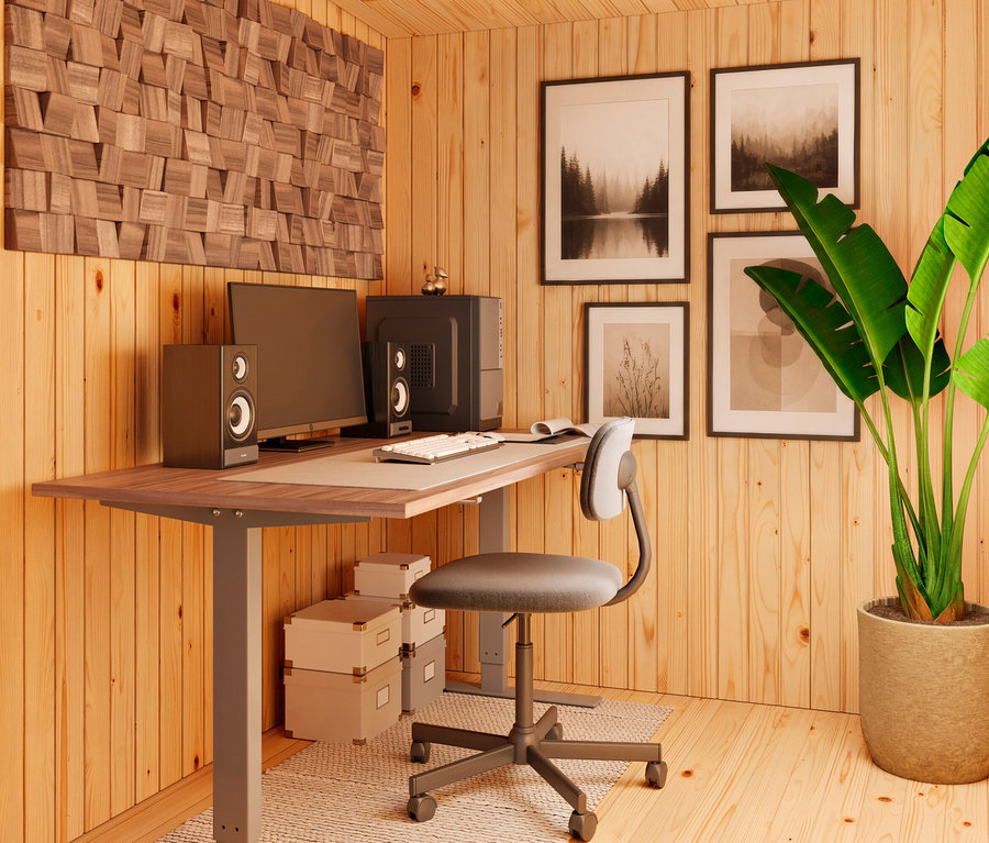 Ein Home-Office-Raum mit hellen Holzpaneelen, einem Schreibtisch mit Computer, Lautsprechern und Tastatur, einem grauen Bürostuhl, drei Aufbewahrungsboxen und einem großen grünen Topfbaum. Eine Holzskulptur und vier gerahmte Bilder schmücken die Wände.