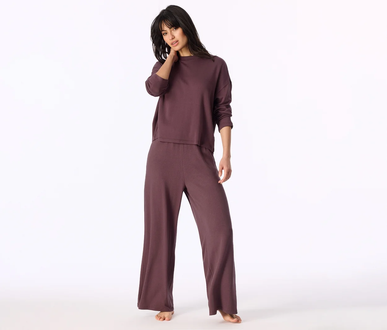 Frau steht in einem dunkelvioletten Langarmshirt und passender weiter Hose, eine Hand am Hals.
