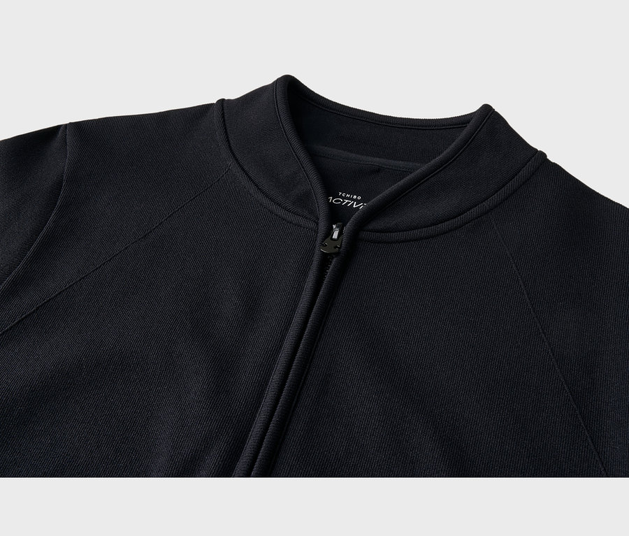 Nahaufnahme einer schwarzen Jacke mit Rippstrickkragen und Reißverschluss, der leicht geöffnet ist. Ein weißes Etikett ist im Nackenbereich sichtbar.