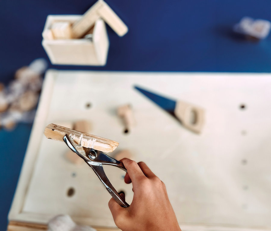 Kinderhand klemmt ein kleines Holzstück mit einer Metallzwinge auf einer small foot Kinderwerkbank, mit einer Spielzeugsäge und Holzklötzen daneben.