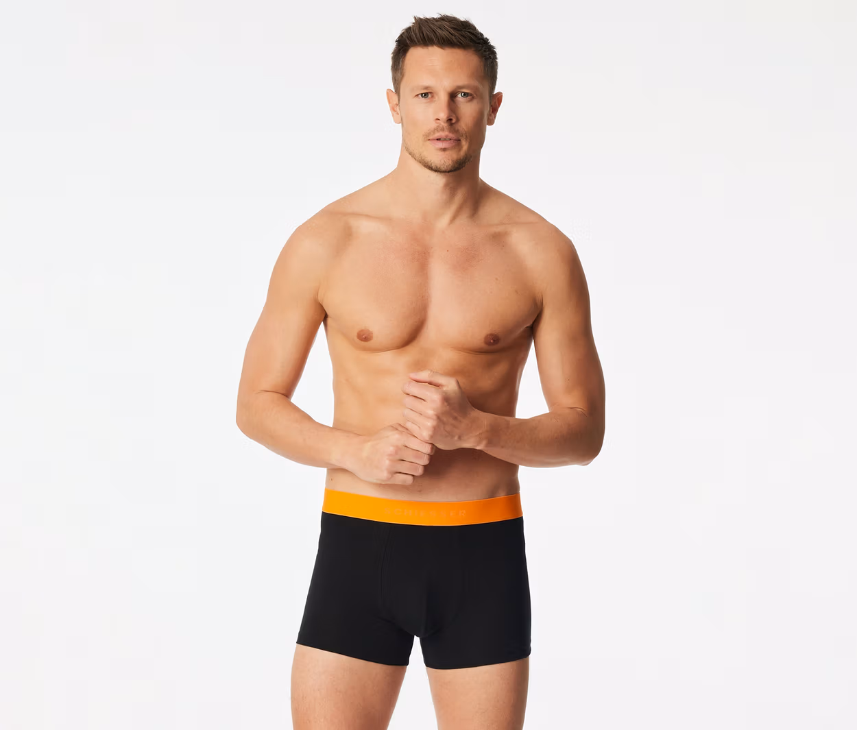 Ein Mann, oberkörperfrei, trägt schwarze Boxershorts mit orangefarbenem Bund. Seine Hände sind vor ihm gefaltet.