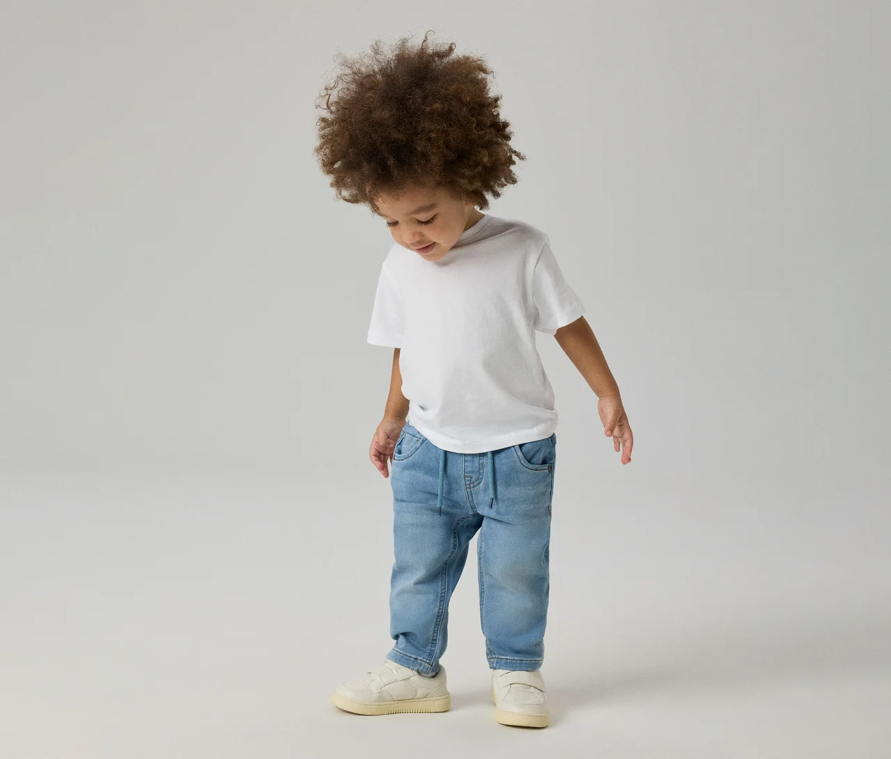 Kleines Kind mit lockigem Haar trägt ein weißes T-Shirt, hellblaue NAME IT Slim Sweat Jeans und helle Sneaker und blickt nach unten.