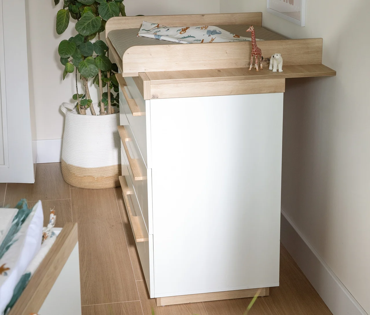 Weißer Wickeltisch mit hellen Holzdetails aus dem Roba 2-teiliges Kinderzimmer-Set »Lumo«. Obenauf liegen eine Wickelauflage mit Tiermuster und Spielzeugtiere. Links steht eine große Pflanze in einem geflochtenen Korb.