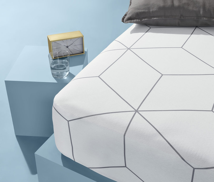 Auf blauem Hintergrund stehen ein Bett mit weißer Bettwäsche mit geometrischem Muster und ein graues Kissen. Neben dem Bett stehen auf einem blauen Tisch eine goldene Uhr und ein Glas Wasser.