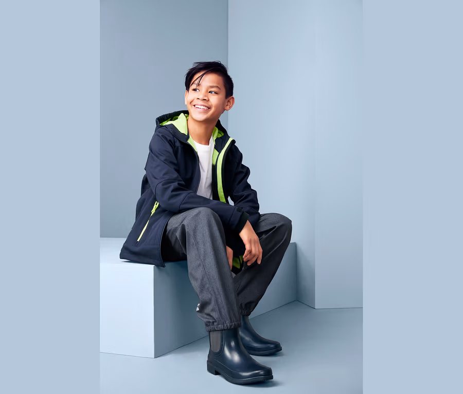 Ein lächelnder Junge sitzt und trägt eine Softshellhose im Jeans-Look, eine Jacke und Gummistiefel.