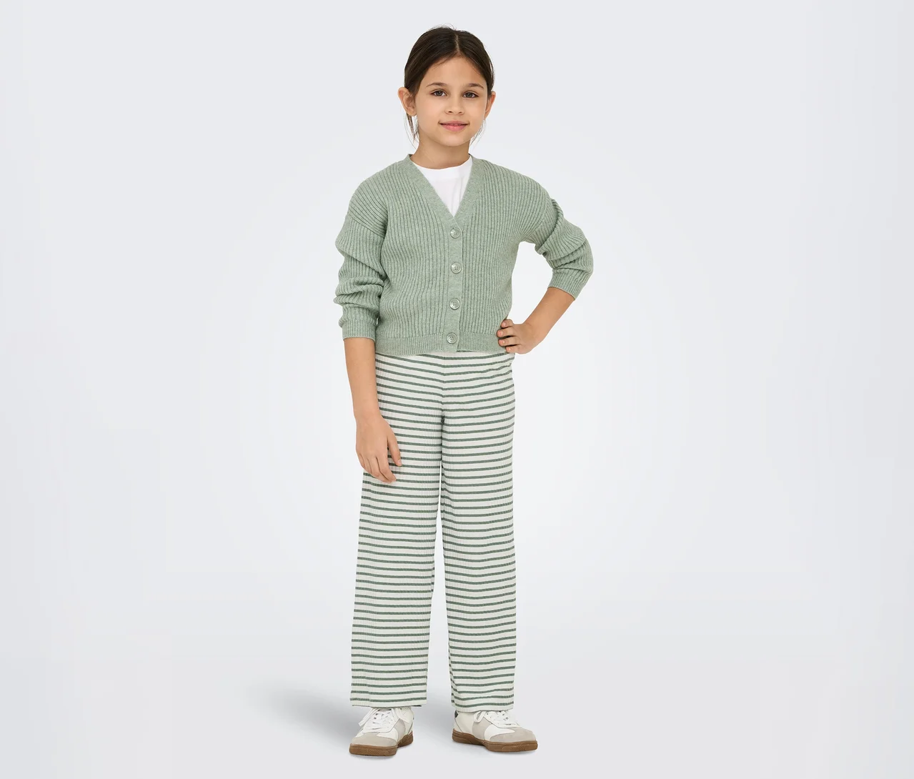Junges Mädchen steht mit den Händen in den Hüften da und trägt eine hellgrüne Strickjacke über einem weißen T-Shirt, gestreifte weite Hosen und helle Turnschuhe.