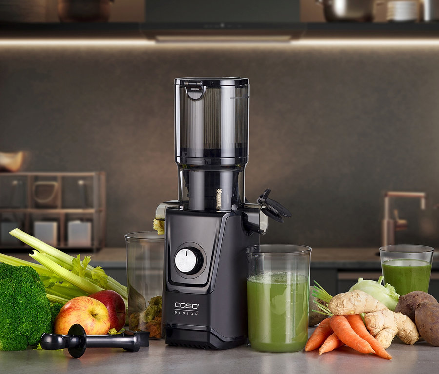 Schwarzer CASO Juice Fit Slow Juicer auf einer Küchenarbeitsplatte, umgeben von frischem Brokkoli, Stangensellerie, Äpfeln, Karotten, Ingwer, Kohlrabi, Roter Bete, einem Glas Trester und zwei Gläsern grünem Saft.