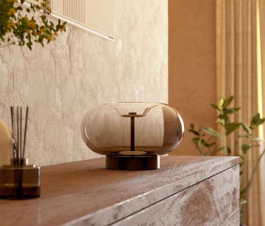 Eine moderne, runde Lampe aus Rauchglas und ein Raumduft-Diffusor stehen auf einer hellbraunen Holzkommode. Im Hintergrund sind eine beigefarbene Wand mit abstrakter Kunst, eine Pflanze und helle Vorhänge zu sehen.