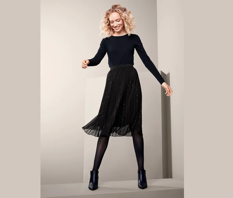 Frau trägt einen Plisseerock, eine schwarze Strumpfhose, schwarze Stiefel und ein schwarzes Langarmshirt.