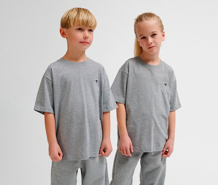 Zwei Kinder, ein Junge und ein Mädchen, stehen nebeneinander und tragen passende graue hummel hmUR loose T-Shirts mit kleinem Bienenlogo und graue Jogginghosen.