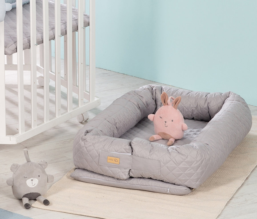 Graue Roba 3-in-1 Babylounge mit Plüschhase auf beigem Teppich neben einem weißen Kinderbett.