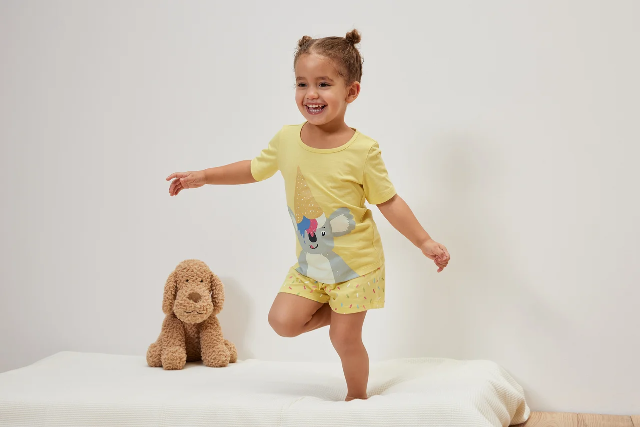 Ein lächelndes kleines Mädchen steht auf einem Bett neben einem Plüschhund und trägt einen gelben Kinder-Shorty-Pyjama, Koala.