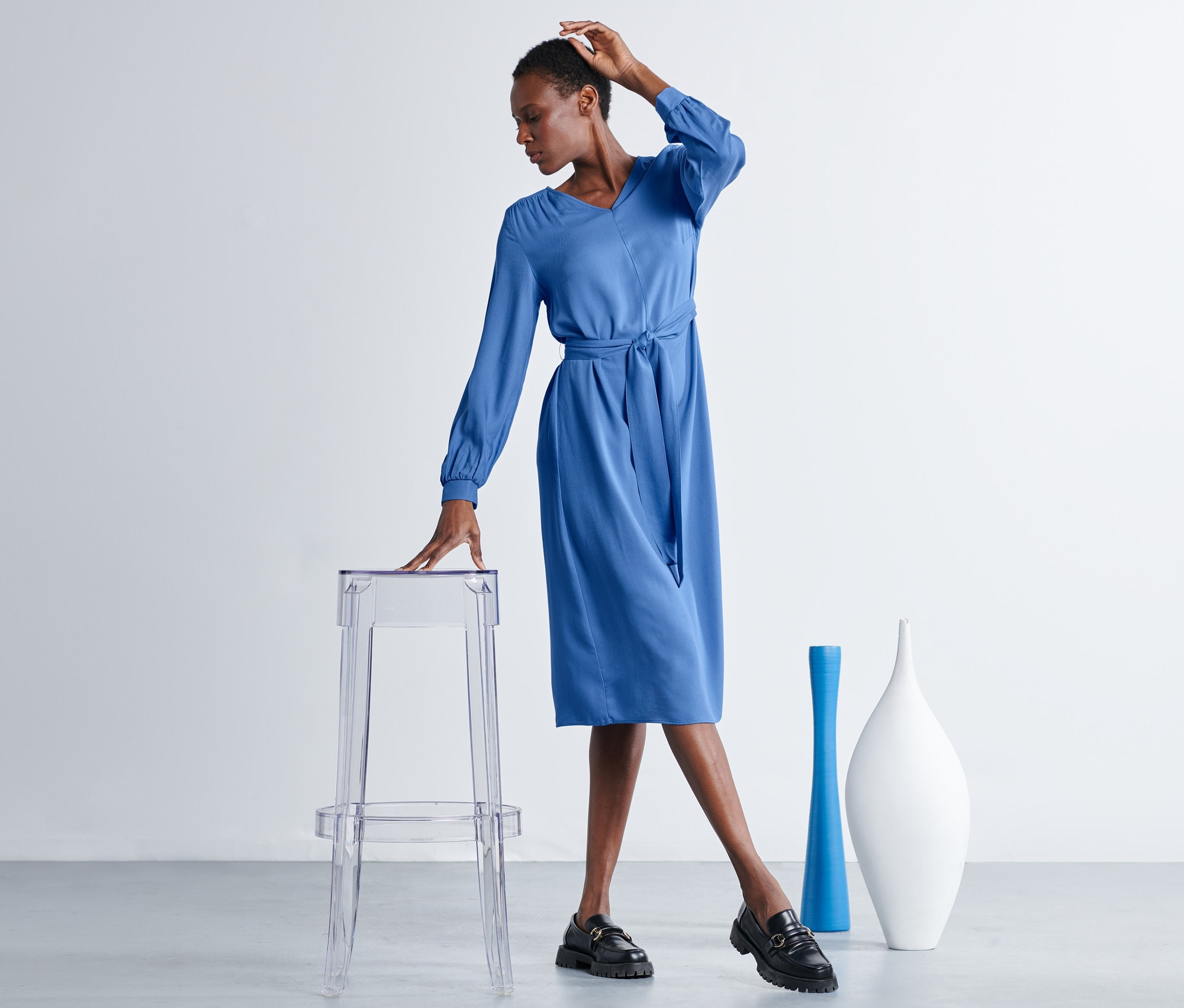 Frau posiert in blauem Midikleid mit Gürtel neben einem transparenten Hocker.