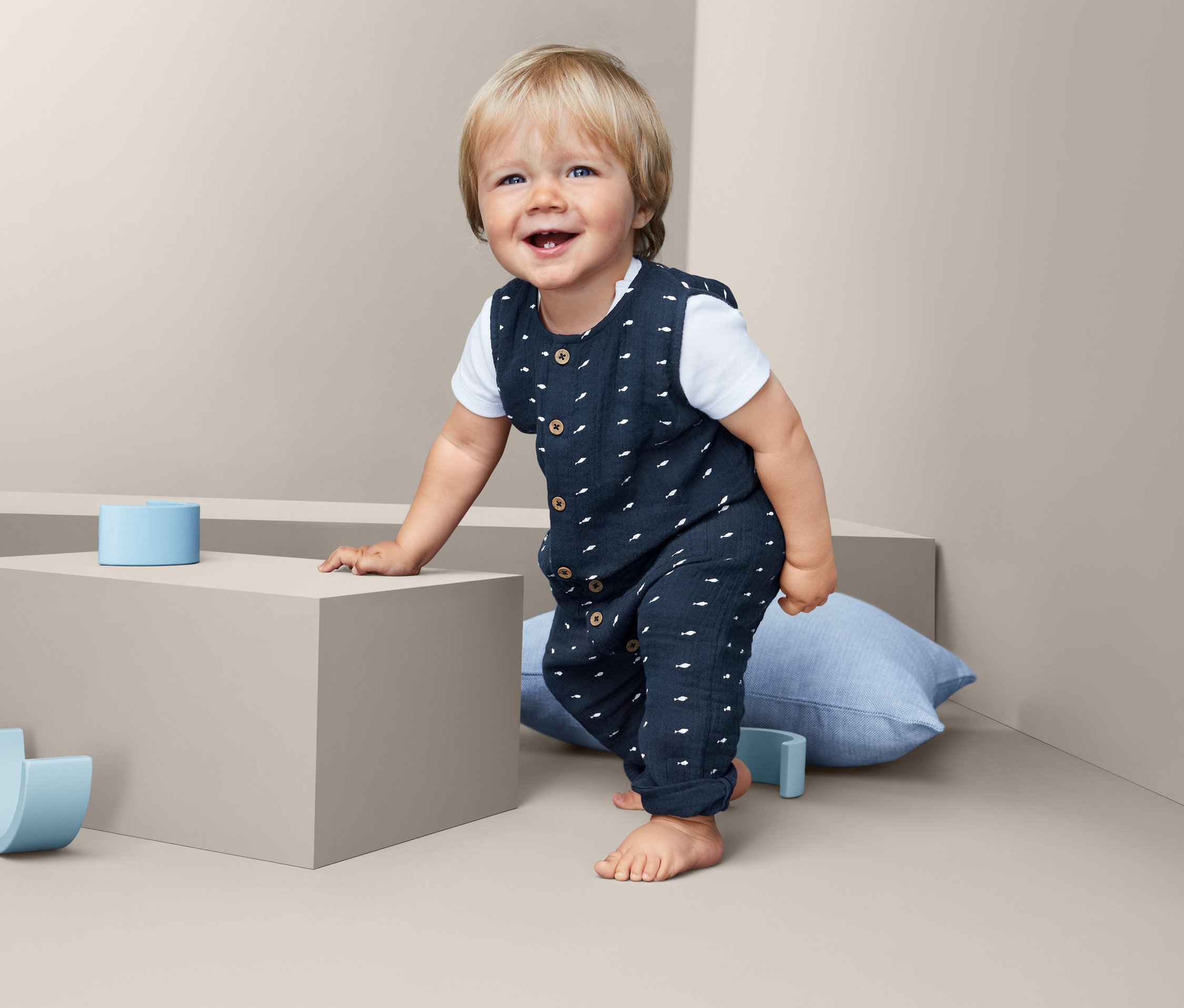 Ein lächelndes Kind steht und lehnt sich an einen grauen Würfel in einem Baby Baumwoll-Strampler.