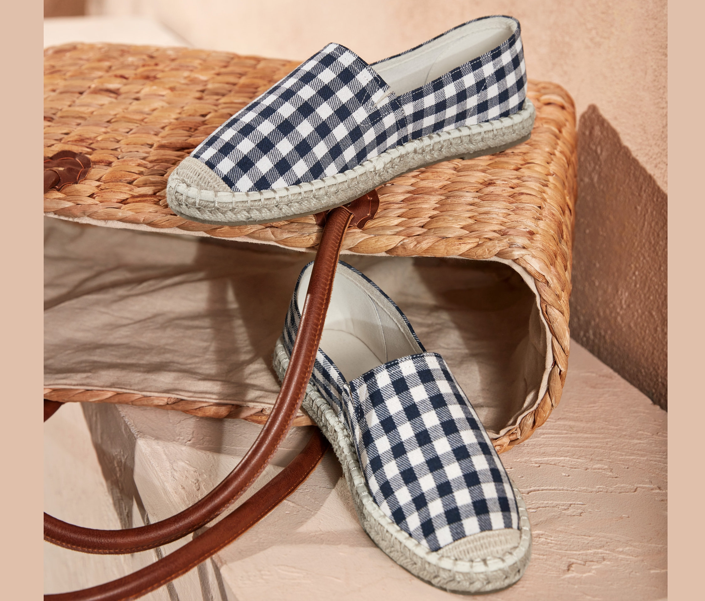 Zwei Espadrilles mit blau-weißem Karomuster liegen auf einer Korbtasche.