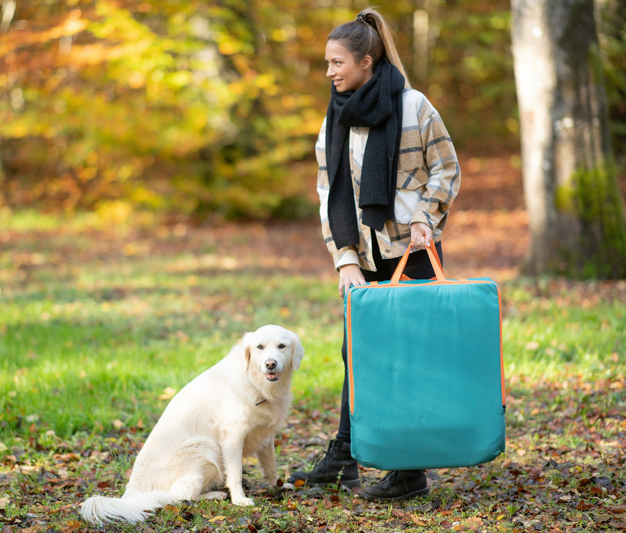 Frau hält eine tragbare Sitzmatte neben einem sitzenden weißen Hund in einem Park.