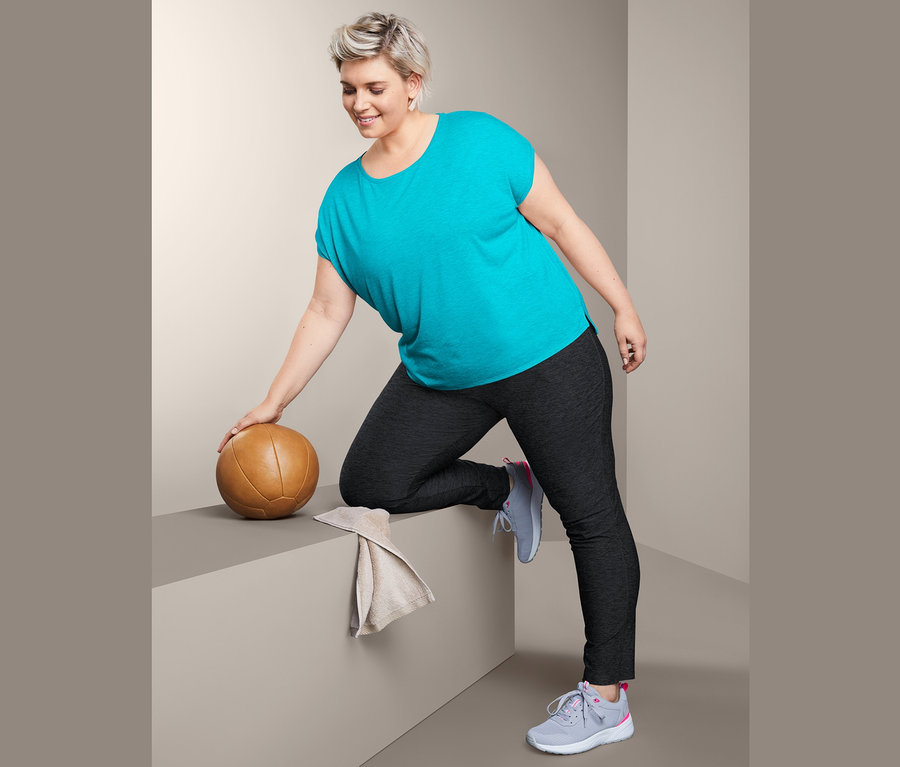 Frau in türkisfarbenem Sportshirt und Trainingshose lehnt sich an einen braunen Medizinball.