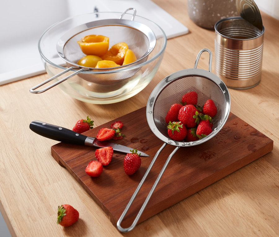 Auf einem Holzbrett liegen ein 2 Edelstahl-Siebe mit Erdbeeren, daneben geschnittene Erdbeeren und ein Messer. Im Hintergrund befinden sich eine Schüssel mit Pfirsichen in einem Sieb und andere Küchenutensilien.