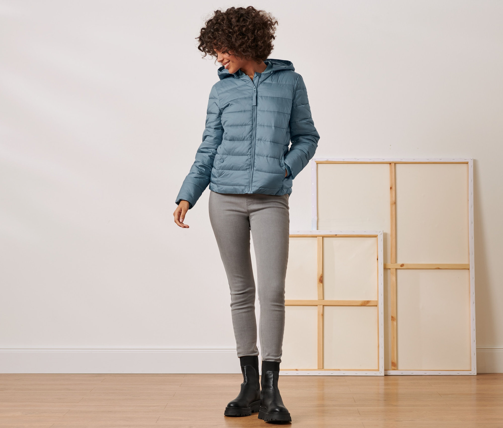 Frau posiert in Steppjacke mit Kapuze, grauen Jeans und schwarzen Stiefeln.