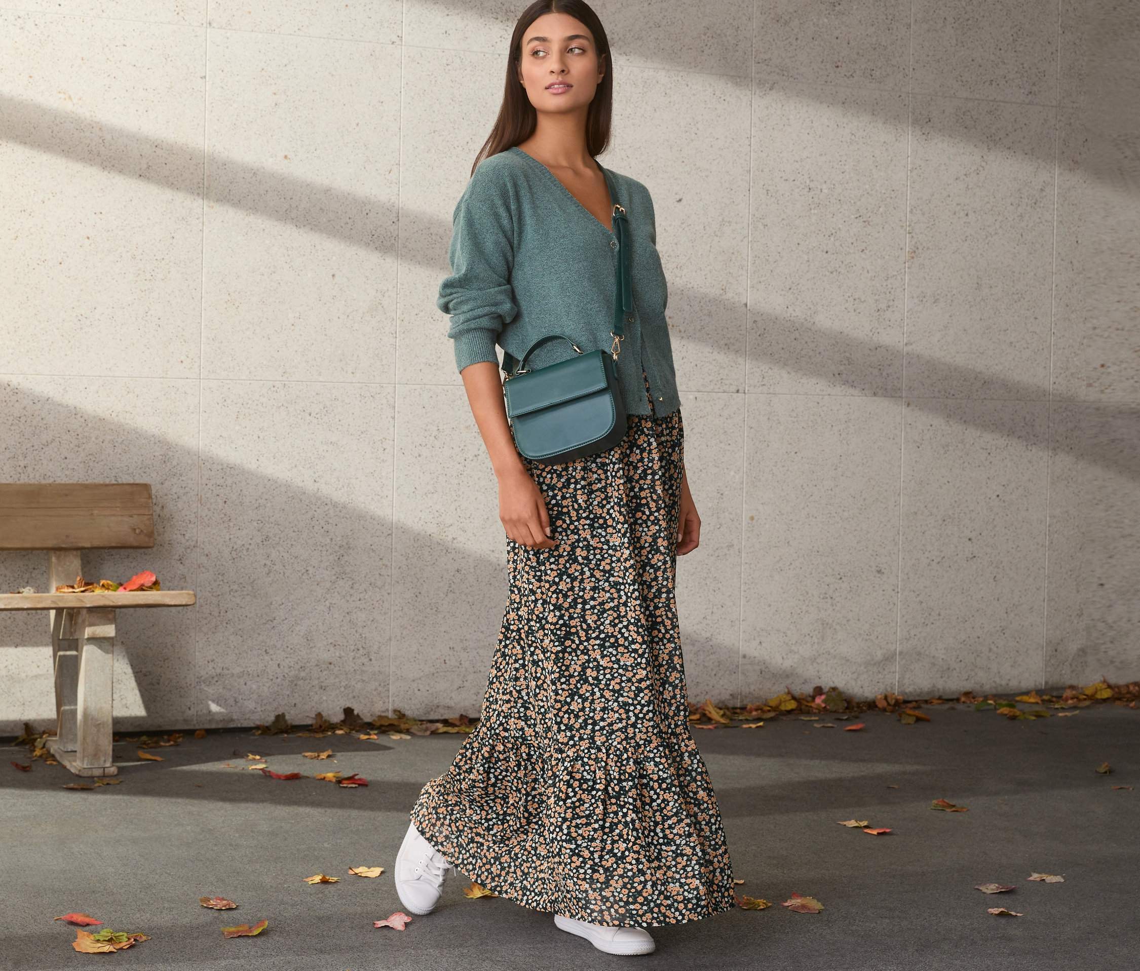 Frau trägt grünen Cardigan und Chiffon-Rock mit Blumenmuster. Sie hat eine grüne Handtasche und weiße Turnschuhe.