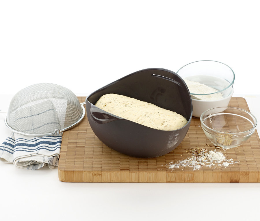 Teig in brauner Lékué Brotback-Set Form auf einem Holzbrett, daneben Sieb, Mehl und Samen.