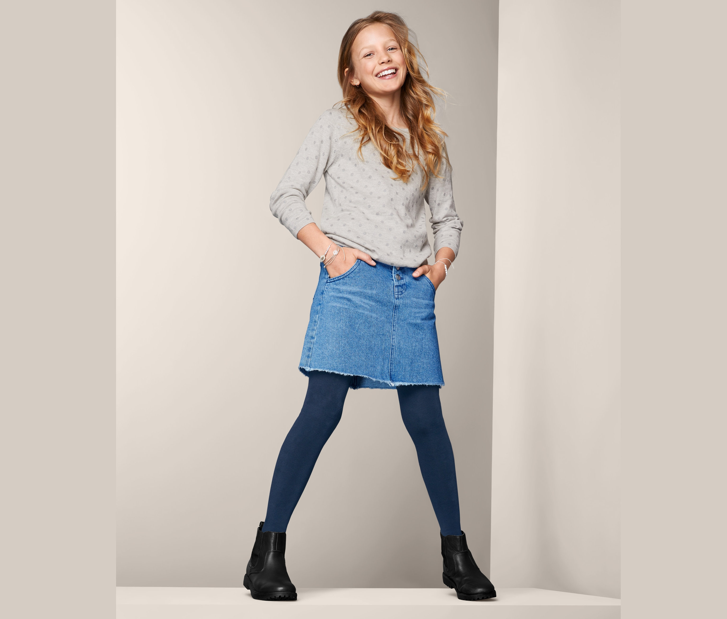 Lächelndes junges Mädchen posiert in grauem Pullover, Jeansrock und blauen 2 Thermostrumpfhosen.
