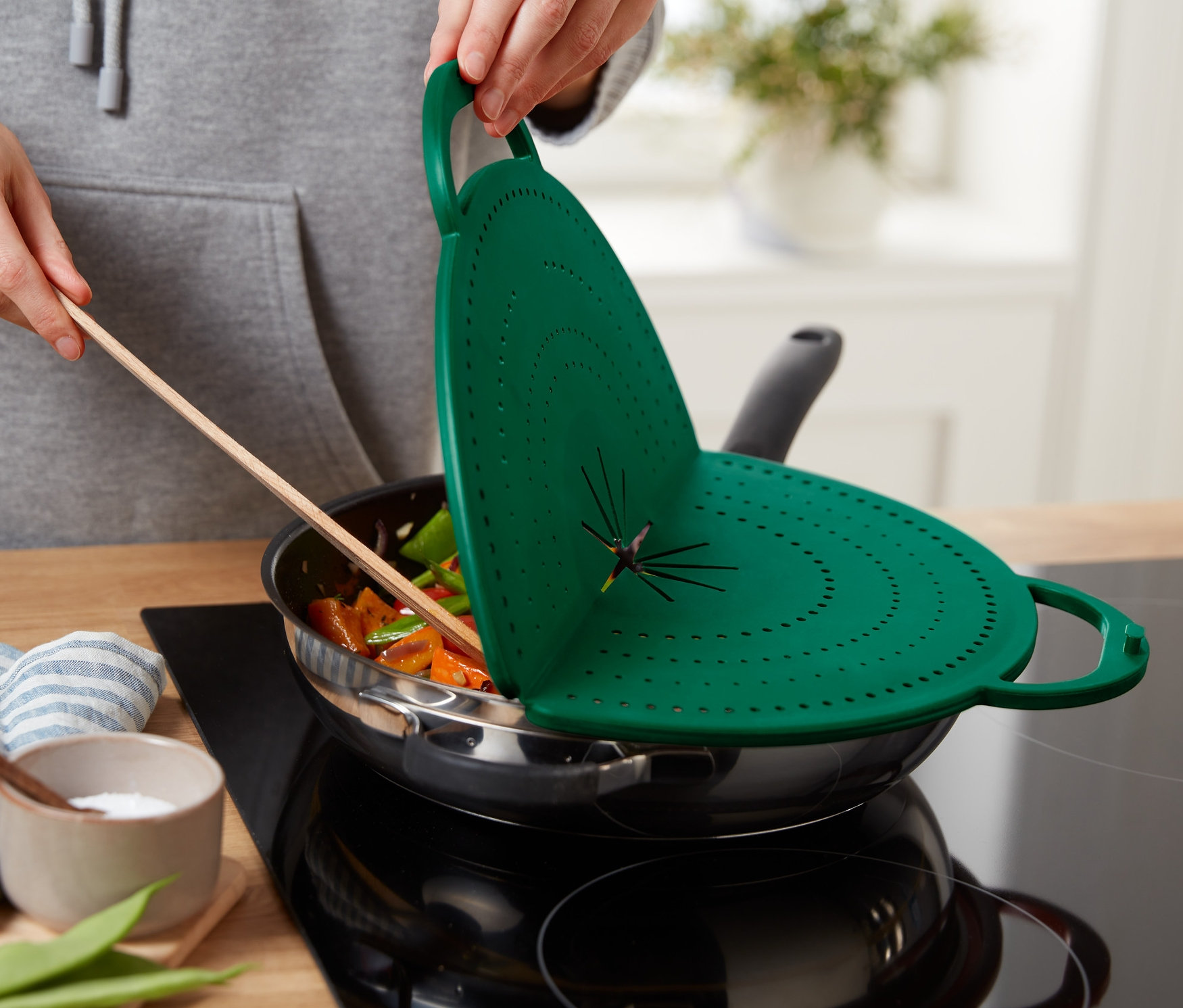 Eine Person hält einen grünen 4-in-1-Pfannen-Spritzschutz über eine Pfanne mit Essen.