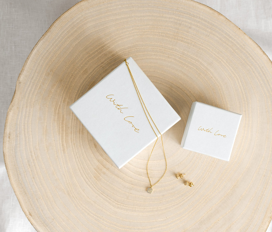 Goldene 585 Gold Kette Zirkonia Cushion und 375 Gold Ohrstecker Zirkonia Cushion auf einem Holzteller neben weißen Schachteln.