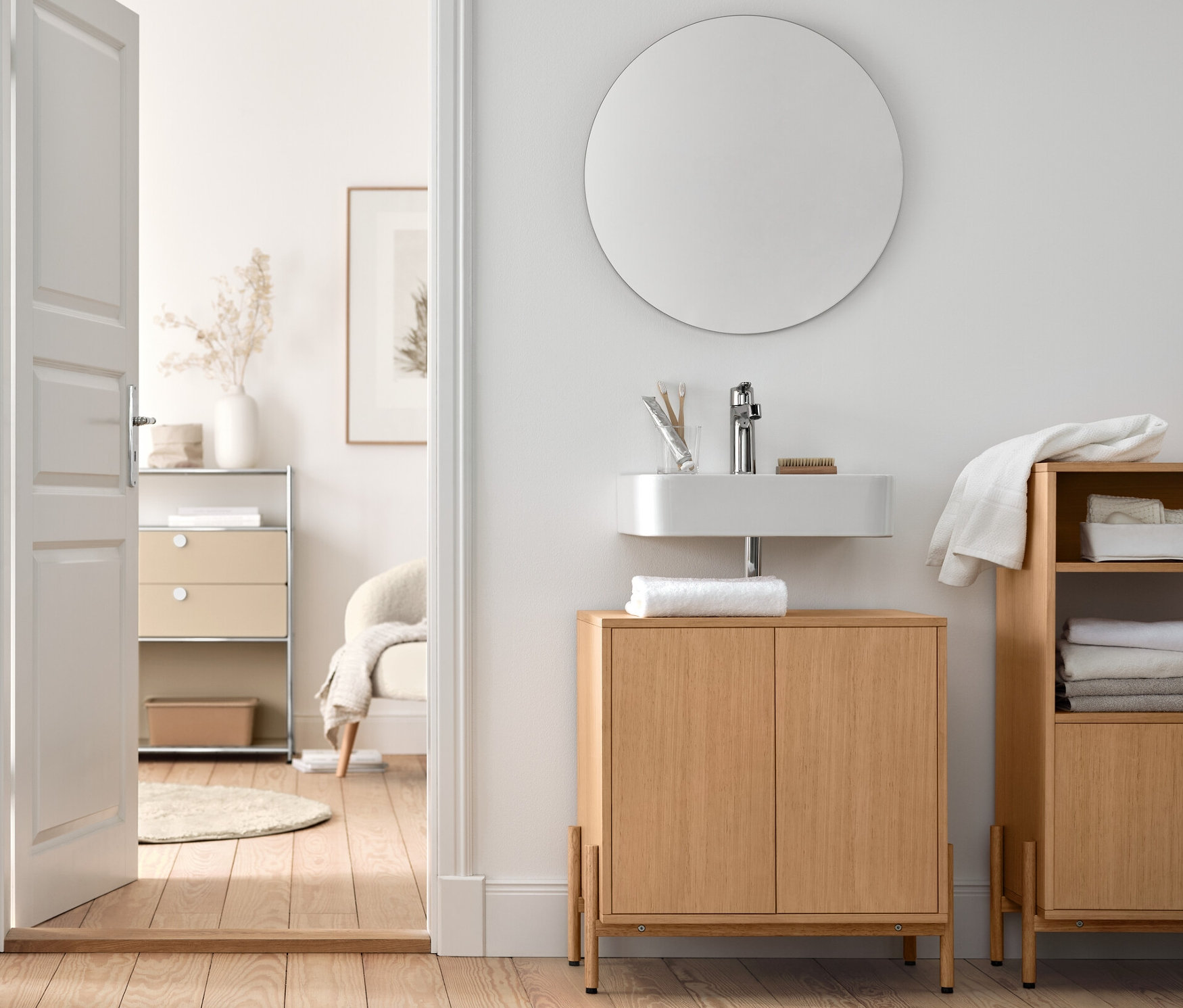 Im Badezimmer stehen ein Waschbeckenunterschrank und ein Badschrank mit Holzfurnier, sowie ein Bouclé-Sessel mit Holzbeinen und ein runder Spiegel »Ameca«.