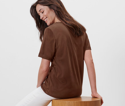 Eine Frau sitzt auf einem Holzhocker und trägt ein braunes T-Shirt mit Stickerei und weiße Hosen.