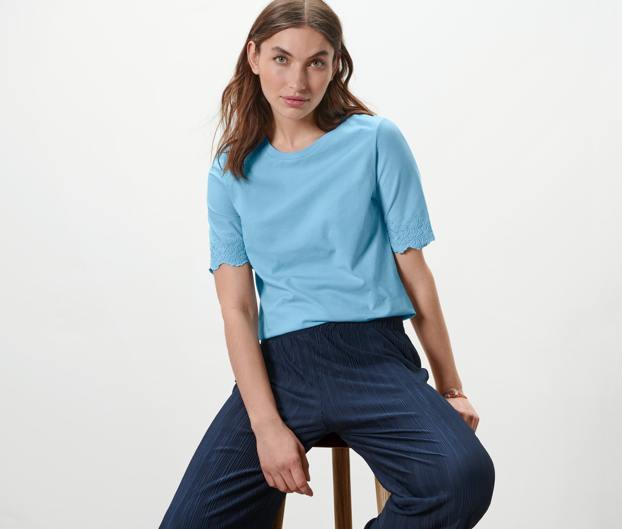 Frau sitzt auf einem Hocker und trägt ein Shirt mit Stickerei.