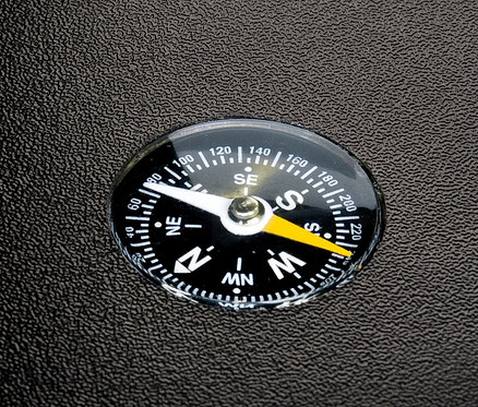 Detailaufnahme eines schwarzen Kompasses mit weißen und gelben Zeigern auf einer schwarzen, strukturierten Oberfläche.