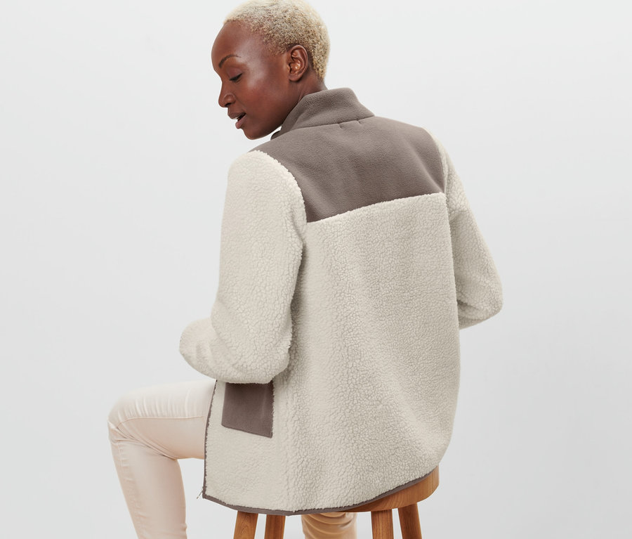 Frau sitzt auf einem Hocker mit dem Rücken zur Kamera und trägt eine beige-braune Teddy-Jacke.