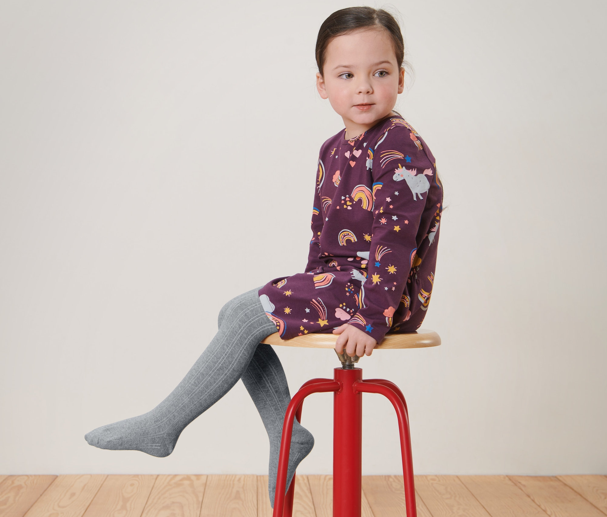 Mädchen sitzt auf einem roten Hocker und trägt ein bedrucktes Kleid und graue 2 Paar Kinder-Strumpfhosen.